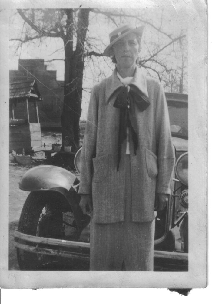 This classically clad lady is Ms. Magdalene Mary Taunton Smith Gresham, sometime in the late 30's, in front of the old Taunton General Store that was built when Red Hill was still called Channahatchee.  

Magdalene Mary Taunton (sometimes Mary Magdalene, but mostly Maggie or Aunt Maggie) was the first child of William Martin and Mary Lee O’Daniel Taunton to survive to adulthood.  Born 29 April 1898, at home in Channahatchee (now Red Hill) in Elmore County, Alabama,  she grew up alongside the Tallassee-Alexander City Road, attending Red Hill School in its earlier incarnations.  A farmer’s daughter, she worked in the home, the yard (swept bare) , in the garden, and in the fields alongside her parents and siblings.  From her mother she received her lifelong love of beautiful flowers.

On graduation from Red Hill School , she went away to “Normal School” to obtain a teaching certificate.  None of us remembers the name or location of the college, or whether she ever taught school.  She was a member of Refuge Missionary Baptist Church, and was active in the doings of the church in the community, sometimes teaching Sunday School classes.

She met and married Simon Wesley Smith, a chiropractor who was born, grew up, trained and practiced in Ohio.  They lived in a beautiful white house across the road and just south from her parents’ home.  She was Dr Smith’s second wife.  Their marriage was short, only about three years , ending with his death in 1929.  She was visited by her stepsons, especially the youngest, Wayne, even after the death of Dr Smith.  Wayne married an Elmore County girl.  He visited last with his children from their home in Arizona.

She worked most of her life at Tallassee Mills, on the second shift, riding Fred Fomby’s blue-and-white “Mill Bus”.  With her sassafras “bresh” and her Bruton Snuff in her mouth, she carried a little brown bag containing her crochet materials to while away the trips to and from work.  
The little brown bag was her constant companion and if she was sitting, she was crocheting, singing under her breath or aloud, “Cheer up my brother, live in the sunshine. Farther along, we’ll understand why”  (I’m certain there were more words to that hymn, but she just repeated these.)  Her doilies and other crocheting projects decorated her home and the homes of her family.  Most of the other women on the bus carried crochet or other homemaking work on the trip to and from Tallassee.

In 1944 she met and married Reuben Peters who lived with her in her little white house in  Red Hill.  Reuben’s son and his family visited from their home in Langdale, Alabama frequently.  The marriage was short, ending in divorce.

She worked constantly in her yard, planting flowers everywhere.  Her favorites were gladiolas, and nieces and nephews were frequently drafted to plant and care for these.  The bulbs were dug up each fall, stored in the garage, and replanted in the spring along with any new ones she had acquired over the winter.  Trading bulbs and plants was another enjoyable activity of the “mill ladies”.  Tall pine trees  flanked her house on the east and south side.  I can remember lying in bed at her house and being lulled to sleep by the wind sighing through the pine needles in that mini-forest.  I also remember being delegated to rake all the pine straw from that mini-forest.  She planted azaleas in the edge of the pines and had a beautiful dogwood blooming just outside her bedroom window,  and a sweet shrub and a gardenia.  Many weddings in the community were enhanced with sprays of blooms from the brides wreath bushes in her front yard.

In 1955, she gave up her job at Tallassee Mills, married the recently widowered husband of her Aunt Maud, Felix Leslie Gresham, and moved with him to his home in Citronelle, in Mobile County.  There she continued her flower garden work, and was active in his church.  She lived there with him until his death in 1963.  She then moved back to her home in Red Hill.

She died of a heart attack about a year later, 15 April 1964, and was buried next to her first husband, S Wesley Smith, in the cemetery at Refuge Baptist Church.

Her former home is now owned by Mrs. Elaine Stewart and has been extensively enlarged and remodeled. 